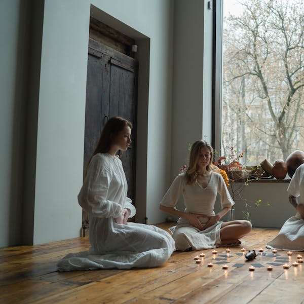 Four women in white dresses meditate with candles on a wooden floor indoors.